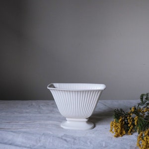 May include: A white, fluted ceramic vase with a square top and pedestal base. The vase has vertical ridges and sits on a white surface. Yellow flowers and green foliage are in the background.