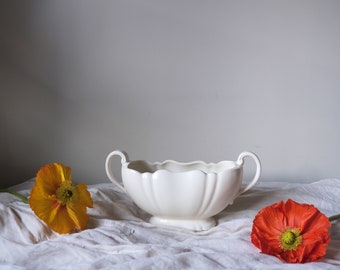May include: A white ceramic bowl with two handles sits on a white fabric surface. Two flowers, one orange and one red, are on either side of the bowl.