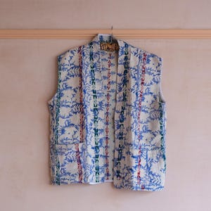 May include: A cream-coloured sleeveless vest featuring a blue floral design and vertical stripes of red, green, and blue embroidery. The vest has two front pockets and hangs on a hanger.
