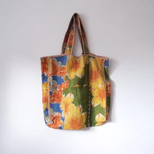 Può includere: Una grande borsa tote colorata con un motivo floreale in tonalità arancio, giallo, verde e blu. La borsa ha un manico in tessuto marrone.