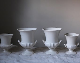 May include: A set of four white ceramic urns with flared rims, fluted bases, and small side handles. The vessels are arranged in a row, featuring a smooth finish and a classic pedestal design that elevates the body of each individual container.