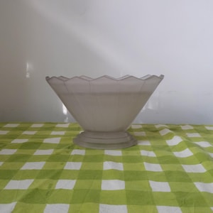 May include: A frosted glass bowl with a scalloped edge and a wide base. The bowl is a light grey colour and is sitting on a green and white checked tablecloth.