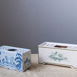 May include: Two rectangular ceramic flower frog vases. The vase on the left has a blue and white design with a squirrel and trees. The vase on the right has a green and white design with two birds.