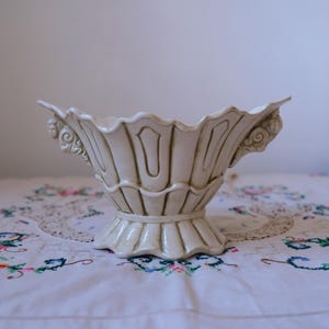 May include: A white ceramic bowl with a decorative, ornate design. The bowl has a scalloped edge and intricate details on the sides. It is sitting on a white tablecloth with a floral pattern.
