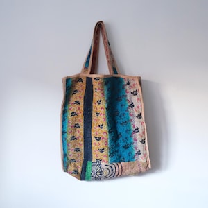 Op de afbeelding: Een kleurrijke patchwork tote bag met blauwe, gele en roze patronen. De tas heeft een lange band en is gemaakt van stof.