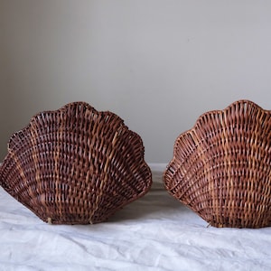 May include: Two brown wicker baskets, each shaped like a scallop shell. The baskets are woven and have a scalloped edge. They are displayed on a white surface against a neutral background.