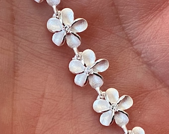 Può includere: Un bracciale argentato con ciondoli a forma di fiore. Ogni ciondolo ha cinque petali e una piccola pietra trasparente al centro. Il bracciale è progettato con una delicata catena che collega i ciondoli floreali.