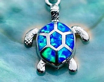 May include: A silver-toned necklace featuring a turtle pendant. The turtle shell is inlaid with iridescent blue and green opal-like material, while the head, legs, and outer shell frame are crafted from polished silver-toned metal on a fine chain.