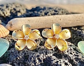Op de afbeelding: Een paar gouden oorknopjes in de vorm van vijfbladige plumeria-bloemen. Elke oorbel heeft een getextureerde, matte gouden afwerking op de bloemblaadjes en een kleine, heldere, fonkelende edelsteen in het midden van de bloem op donker gesteente.