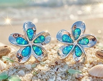 Può includere: Un paio di orecchini a lobo color argento a forma di fiore con cinque petali ciascuno. Il centro di ogni petalo è intarsiato con una vibrante pietra di opale blu iridescente che mostra bagliori di luce verde e ciano sul metallo lucido.