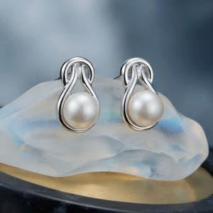 May include: A pair of silver stud earrings featuring a polished metallic loop design that cradles a single round white pearl. The earrings have a smooth, reflective finish and a secure post backing, showcasing a clean and symmetrical aesthetic.