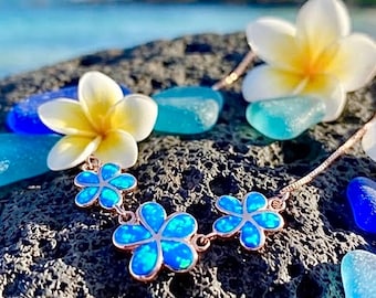Op de afbeelding: Een ketting met drie bloemvormige bedels met helderblauwe iriserende inlays in een metalen frame. De ketting wordt getoond naast witte en gele plumeria-bloesems en verschillende gladde, doorschijnende blauwe stukjes zeeglas.