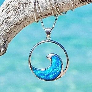 May include: A silver necklace featuring a circular pendant with a vibrant blue opal inlay shaped like a crashing ocean wave. The pendant hangs from a delicate silver box chain, showcasing a polished metallic finish and a bright, iridescent blue color.