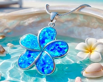 Op de afbeelding: Een zilveren hanger in de vorm van een vijfpuntige plumeria-bloem, voorzien van een levendige blauwe iriserende inleg. De hanger hangt aan een fijne zilveren ketting en rust in een grote, gladde, parelmoerkleurige schelp gevuld met helder water.