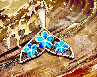 May include: A silver pendant necklace with a tail-shaped design, adorned with three blue and turquoise flower-shaped opals. The pendant is suspended from a fine silver chain, set against a textured, iridescent backdrop.