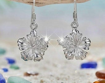 May include: A pair of silver-toned drop earrings shaped like hibiscus flowers. Each earring features a central stamen accented with small, clear, sparkling stones. The earrings hang from simple silver-toned hooks and have a polished metallic finish.