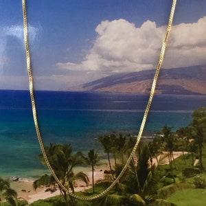 May include: A gold chain necklace with a box chain design. The chain is laying on a background of a beach scene with palm trees and blue water.