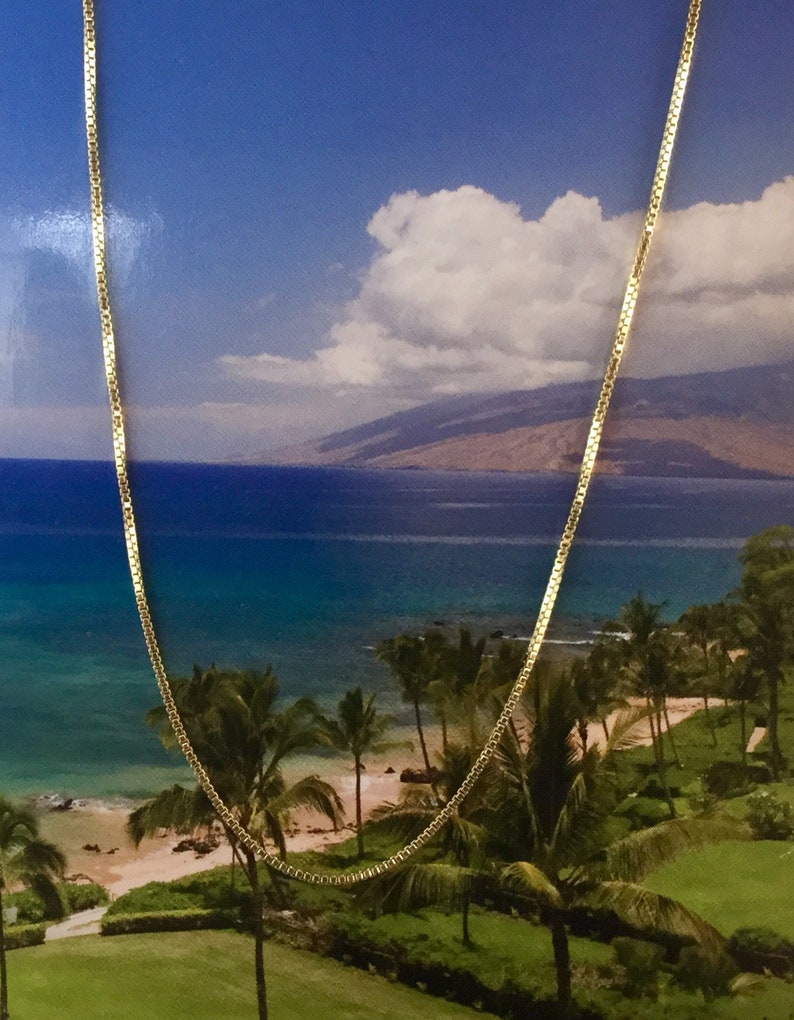 May include: A gold chain necklace with a box chain design. The chain is laying on a background of a beach scene with palm trees and blue water.