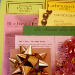 May include: A close-up of a pink, green, and yellow gift bag with a gold bow. The bag is filled with various brochures and pamphlets about jewelry, including Hawaiian Peridot Jewelry, Labradorite Jewelry, Caring for Pearls, and The Hawaiian Shape Cut. The text "Blue Hawaii" is visible on one of the brochures.