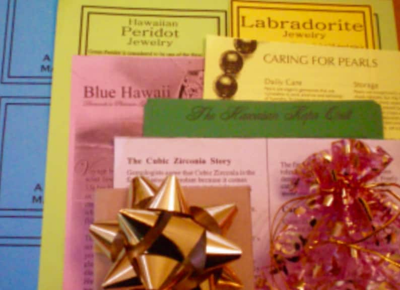 May include: A close-up of a pink, green, and yellow gift bag with a gold bow. The bag is filled with various brochures and pamphlets about jewelry, including Hawaiian Peridot Jewelry, Labradorite Jewelry, Caring for Pearls, and The Hawaiian Shape Cut. The text "Blue Hawaii" is visible on one of the brochures.