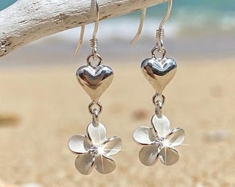 May include: A pair of silver drop earrings featuring a polished heart charm suspended above a matte-finish five-petal flower charm. Each flower centre is accented with a small, clear, sparkling stone, and the earrings hang from silver hooks.