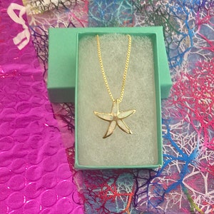 May include: A gold chain necklace with a white opal starfish pendant in a light green gift box. The box is on a pink and white patterned background.