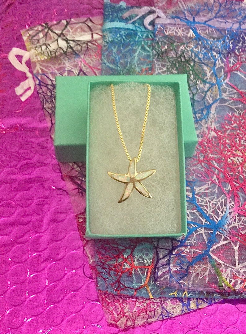 May include: A gold chain necklace with a white opal starfish pendant in a light green gift box. The box is on a pink and white patterned background.
