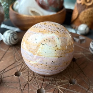 May include: A polished stone sphere with a mix of beige, pink, and brown hues. The orb is set on a wooden surface with a geometric pattern. Other decorative items, including a wooden owl and other stone spheres, are visible in the background.