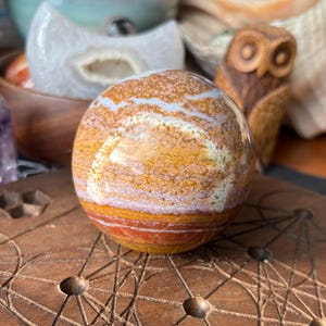 May include: A polished gemstone sphere with bands of orange, brown, and white. The sphere is resting on a wooden surface with a geometric design. Other decorative items are in the background, including a carved wooden owl.