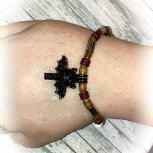 Peut inclure: Un bracelet en perles brunes et bordeaux avec un charme en métal argenté en forme de chauve-souris.