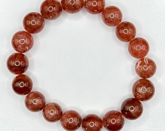 May include: A bracelet made of reddish-brown, round beads. The beads have a translucent quality and are strung together on an elastic band. The bracelet is displayed on a white, circular surface against a black background.
