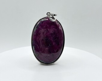 Könnte beinhalten: Ovaler Anhänger mit einem tiefvioletten Edelstein. Der Stein ist in einen silberfarbenen Metallrahmen gefasst und hat oben eine kleine Schlaufe für eine Kette. Der Anhänger wird auf einer weißen Oberfläche präsentiert.