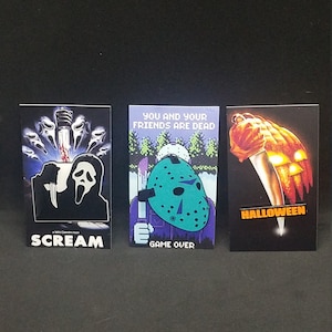 May include: Three horror movie themed pins. The first pin is black with a white outline of a ghost face from the movie Scream. The second pin is purple with a green hockey mask from the movie Friday the 13th. The third pin is black with an orange knife and a pumpkin from the movie Halloween. The text on the pins reads "Scream", "You and your friends are dead, Game Over", and "Halloween".