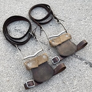 May include: Two pairs of brown leather stirrups with silver metal buckles and straps. The stirrups have a worn, distressed look and are marked with the initials "JV".