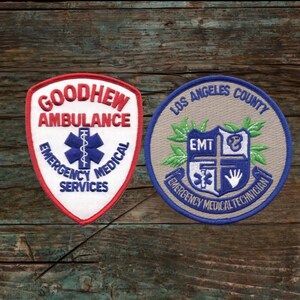 Peut inclure: Deux patchs brodés sur un fond en bois. Le patch de gauche est rouge, blanc et bleu avec le texte "Goodhew Ambulance Emergency Medical Services". Le patch de droite est beige avec le texte "Los Angeles County EMT Emergency Medical Technician".