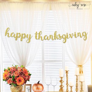 May include: A gold glitter "happy thanksgiving" banner hangs in front of a window with white curtains. A floral arrangement with orange and red flowers sits in a gold vase. A pumpkin with "give thanks" is also visible.