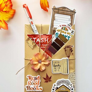 A fall-themed Blind Date with a Book bundle wrapped in kraft paper and twine, decorated with autumn stickers like a pumpkin and books, and includes a highlighter, tea, book tabs, and a bookmark, adorned with a flower, giving off cozy fall vibes.