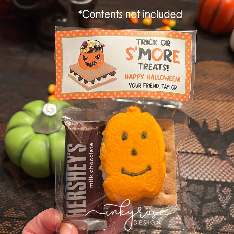 May include: A clear plastic bag filled with Halloween treats. The bag contains a chocolate bar, a graham cracker, and a pumpkin-shaped marshmallow. A tag attached to the bag reads "Trick or S'more Treats! Happy Halloween! Your Friend, Taylor".