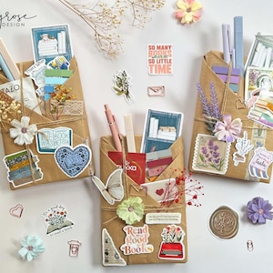 May include: Three brown paper pocket organizers decorated with stickers, pens, and dried flowers. The organizers are tied with twine and feature stickers with book-related quotes and images. One sticker reads "So many books so little time."