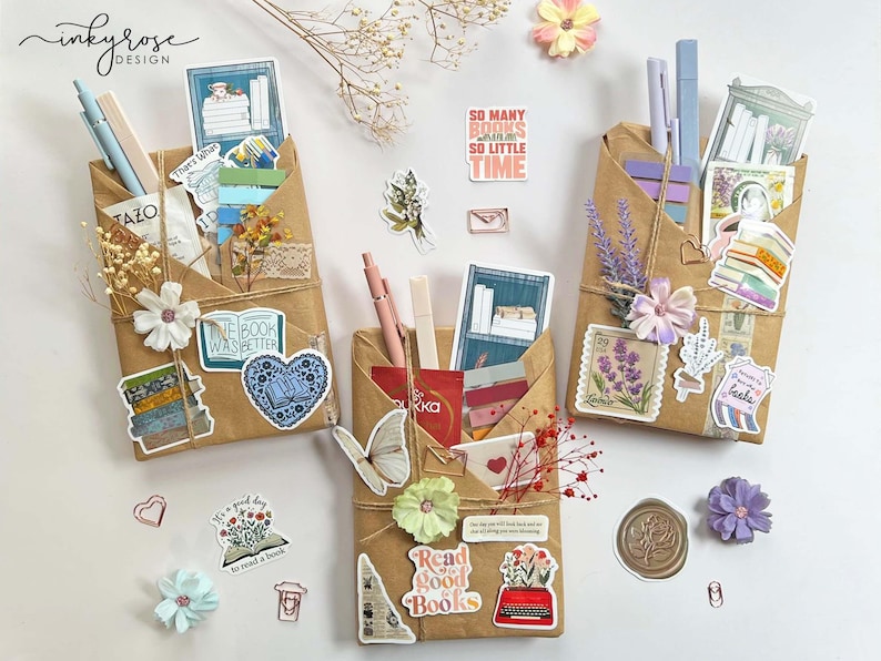 May include: Three brown paper pocket organizers decorated with stickers, pens, and dried flowers. The organizers are tied with twine and feature stickers with book-related quotes and images. One sticker reads "So many books so little time."