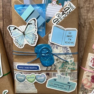 May include: A brown paper package tied with blue twine, sealed with a blue wax stamp. Adorned with stickers of a butterfly, a book, and text that reads "Go Away, I'm Reading" and "Love Book Club". Includes blue sticky notes and a tea bag.