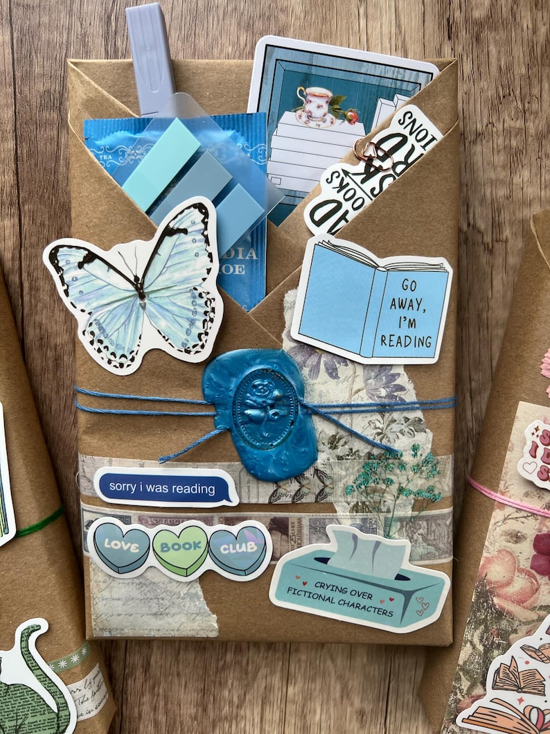 May include: A brown paper package tied with blue twine, sealed with a blue wax stamp. Adorned with stickers of a butterfly, a book, and text that reads "Go Away, I'm Reading" and "Love Book Club". Includes blue sticky notes and a tea bag.