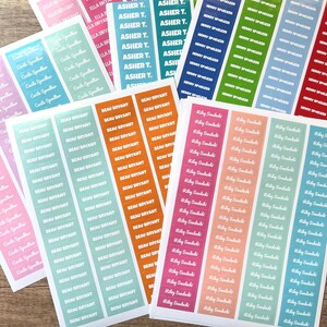 May include: Sheets of colorful name labels with names printed in white. The labels are in various colors, including pink, blue, green, orange, and red. The names are printed vertically.