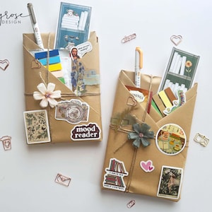May include: Two brown paper-wrapped packages, decorated with stickers, pens, and twine. One has a white flower and a "mood reader" sticker. The other has a blue-green flower and a "I have no shelf control" sticker. Stationery and decorative items.
