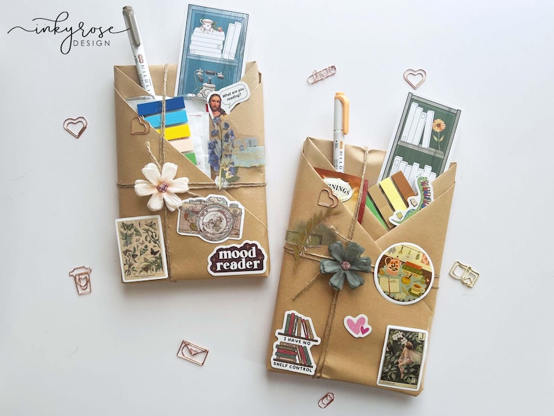 May include: Two brown paper-wrapped packages, decorated with stickers, pens, and twine. One has a white flower and a "mood reader" sticker. The other has a blue-green flower and a "I have no shelf control" sticker. Stationery and decorative items.