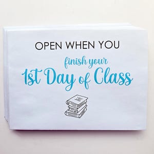 May include: A stack of white rectangular envelopes featuring the black text OPEN WHEN YOU and the blue script text finish your 1st Day of Class, accompanied by a small black line drawing of a stack of three books centered at the bottom.