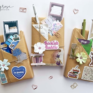 May include: Three brown paper pocket organizers decorated with stickers, twine, and paper flowers. Each pocket holds pens, tea packets, and decorative stickers with book-themed designs. The organizers are arranged on a white surface.