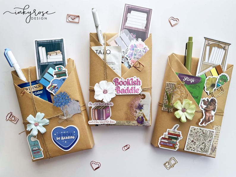 May include: Three brown paper pocket organizers decorated with stickers, twine, and paper flowers. Each pocket holds pens, tea packets, and decorative stickers with book-themed designs. The organizers are arranged on a white surface.