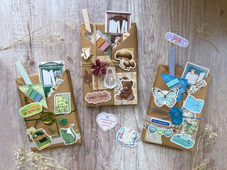May include: Three brown paper packages decorated with stickers, twine, and paper clips. The packages are adorned with stickers of books, a cat, a teddy bear, a butterfly, and the words "Books + Coffee" and "Sorry I was reading".