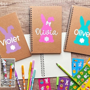May include: Three brown spiral notebooks with bunny designs and names "Violet", "Olivia", and "Oliver" on the covers. The notebooks are on a wooden surface with stickers and pens.